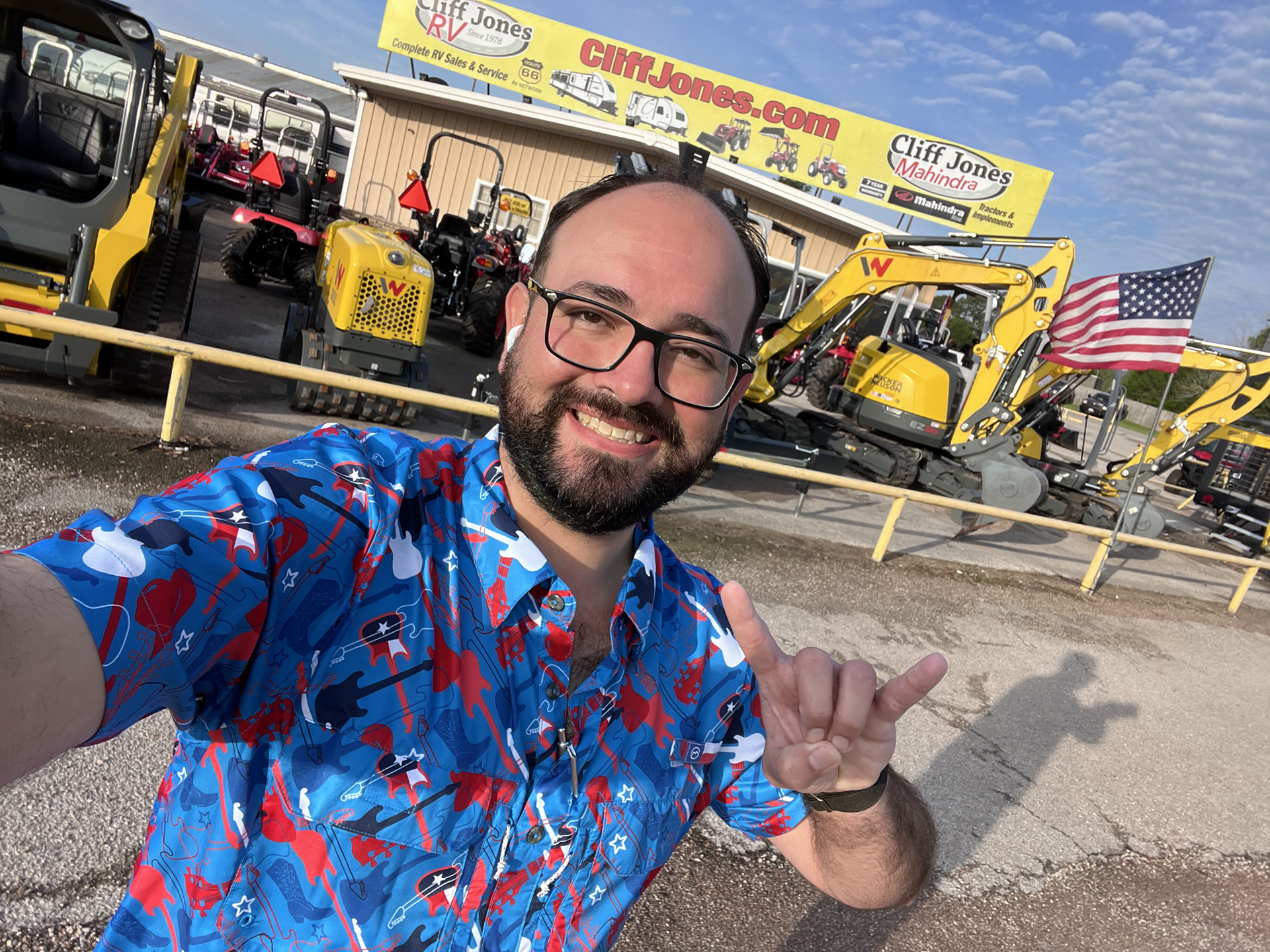 A man with a beard and glasses is standing in front of a construction site. He is wearing a blue shirt and making a peace sign with his hand. In the background, there is a yellow bulldozer parked in a parking lot. The man appears to be smiling. The construction site includes various machinery vehicles and a flag. The man''s face is the focus of the image, with his glasses adding to his distinctive appearance. The overall scene conveys a sense of industrial work and positivity.